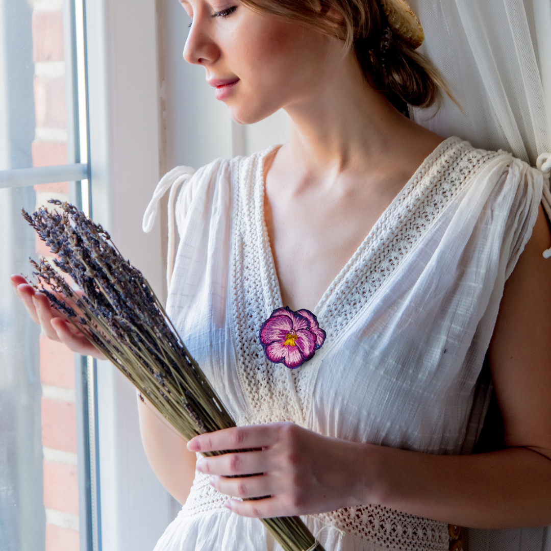 A hand-embroidered pansy brooch, delicate and artistic accessory showcasing intricate craftmanship is adorned by a beautiful girl in a white lacy dress holding a bunch of lavender flowers and admiring natures beauty lovingly