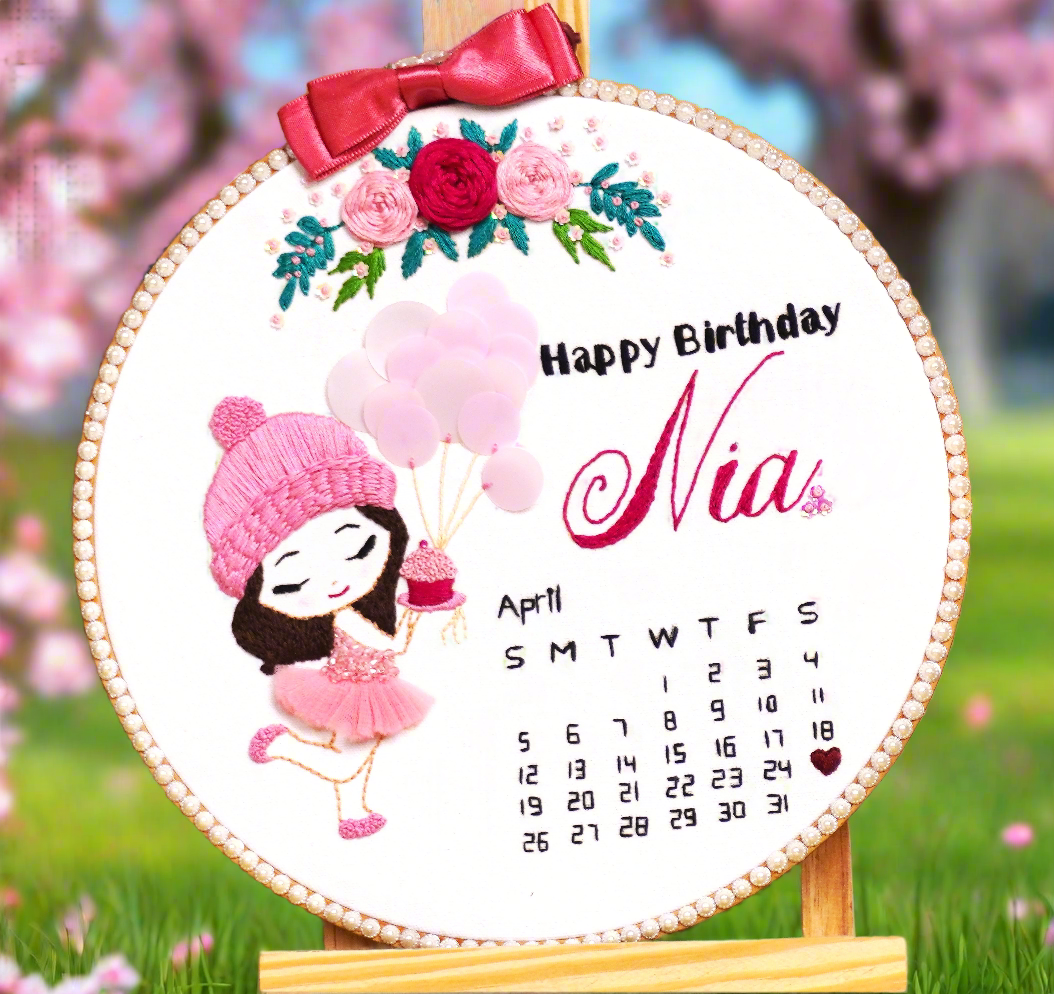 birthday embroidery hoop art with little girl caricature cartoon in pink dress holding pink ballons and a pink cup cake. happy birthday lettering in black color with name embroidery in pink color and pink bow on the top of the embroidery hoop with pink roses and green leaves