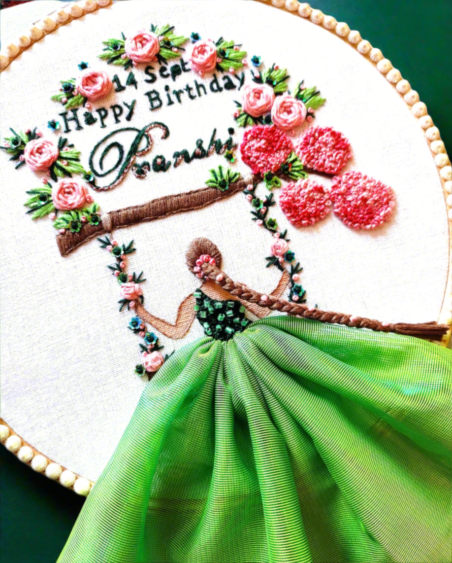 embroidery hoop art with a little girl wearing green color dresses sitting on a swing hanging from cherry blossom tree branch with roses and embroidered name lettering and happy birthday lettering with date and month details