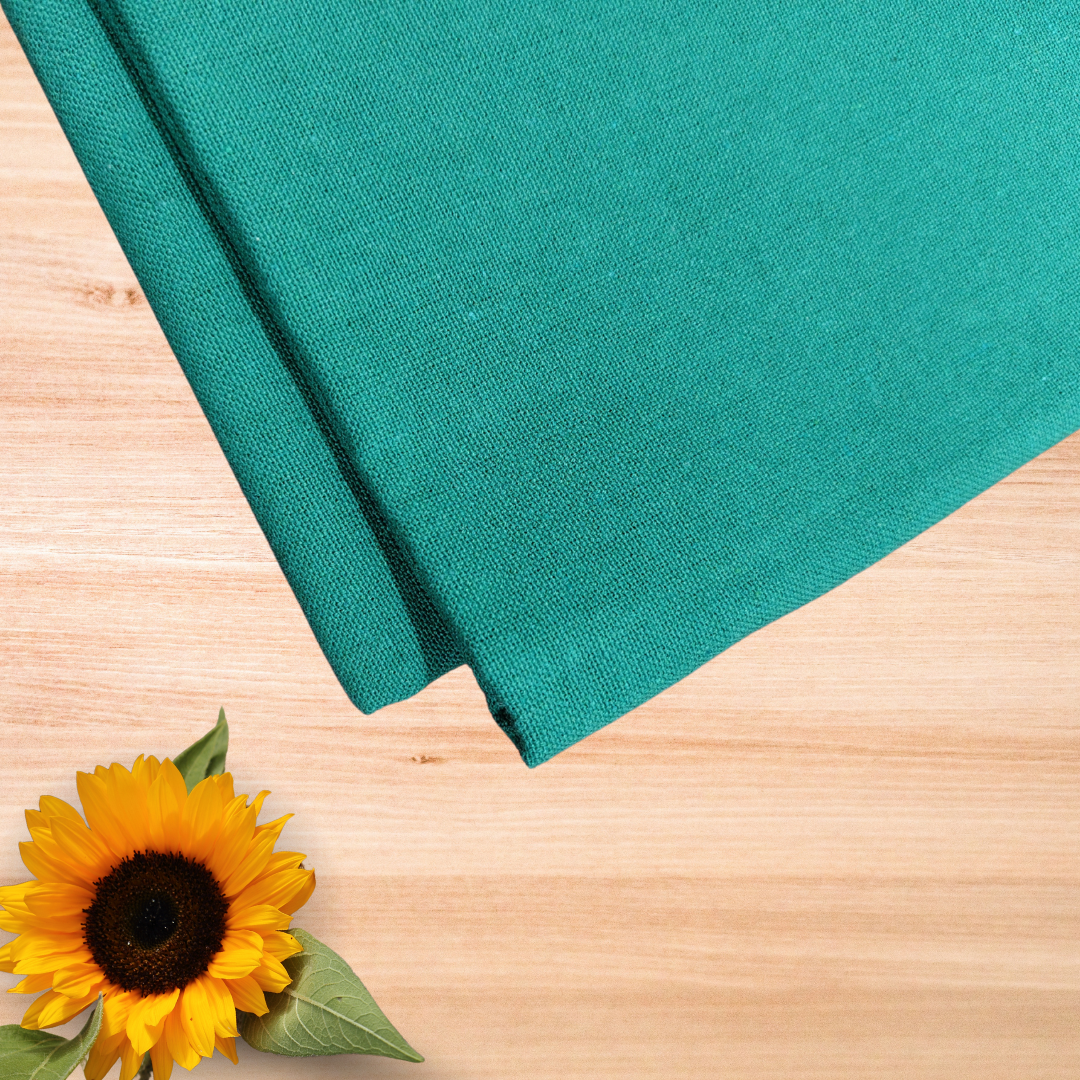 top view of ideal casement embroidery fabric in teal green color placed on a light brown wooden table with a sunflower and 3 leaves