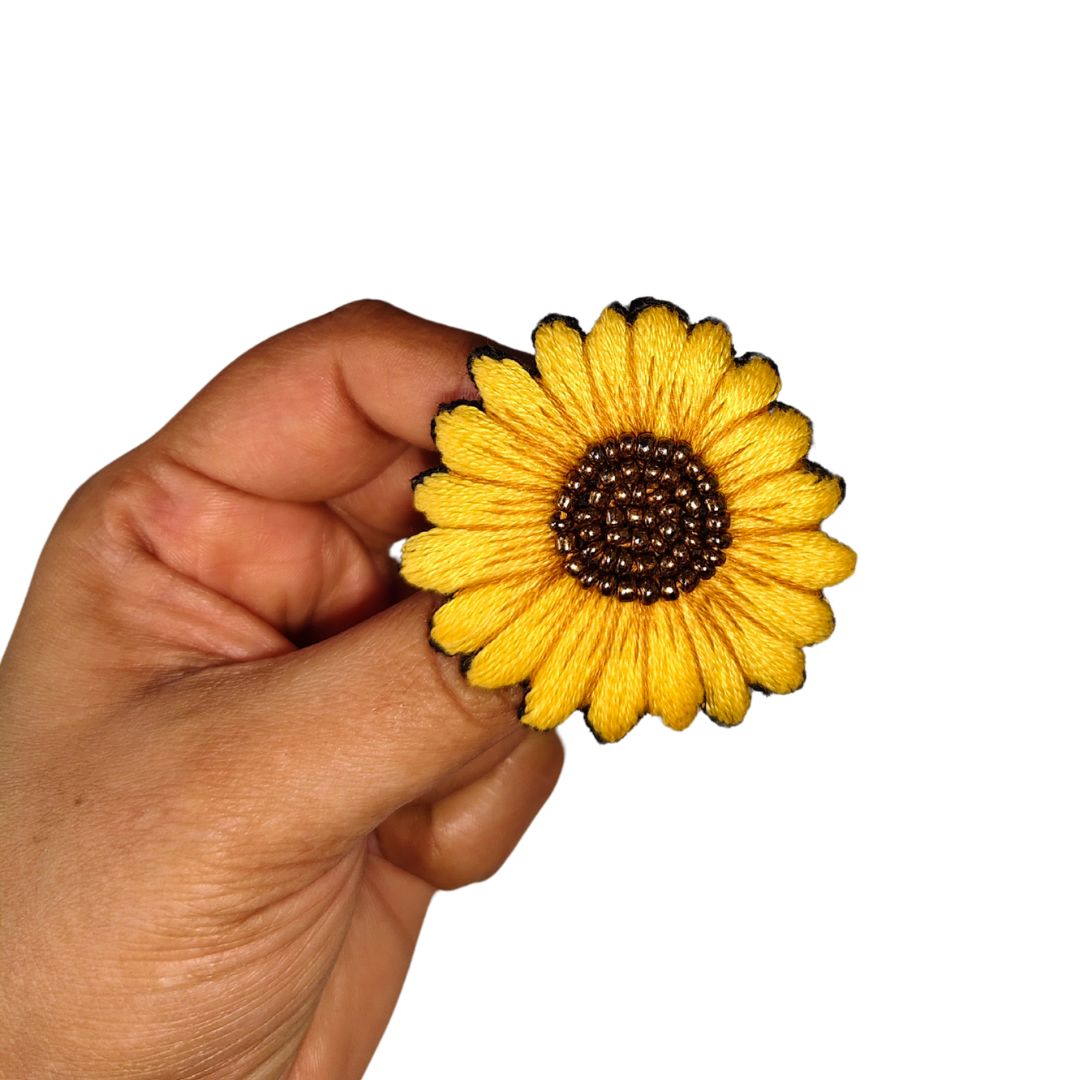 a woman's hand holding a thready sunflower brooch with beads with fingertips in front white background