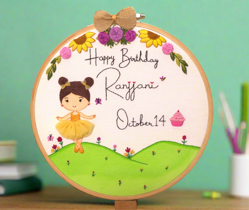 A beautiful birthday embroidery hoop art customized with name and date and happy birthday lettering featuring a cute little girl cartoon caricature, dressed in cute orange frock with cute space buns hair embroidery adorned with 2 hair bows, standing on a hand painted hill, with beautiful flowers in yellow and purple colors, tiny colorful flowers with 2 butterflies in pink color, bee and a cute little embroidered cup cake. the hoop art is further decorated with a jute bow on top.