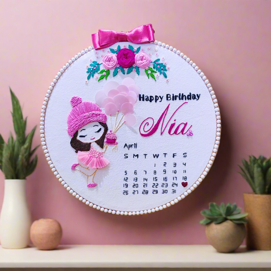 birthday embroidery hoop art with little girl caricature cartoon in pink dress holding pink ballons and a pink cup cake. happy birthday lettering in black colour with name embroidery in pink color and pink bow on the top of the embroidery hoop with pink roses and green leaves