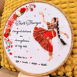 A circular embroidered cross-stitch piece on a white fabric background, featuring a dancing woman in red and brown traditional attire, floral embroidery on the left, and personalized text reading 'Dear Dhanya Congratulations on Aranganrom 25 July 2026 God Bless You Aja Dodda Arjun Nidhi'. The hoop is displayed against a yellow textured pom-pom cushion.