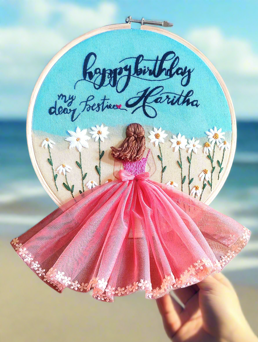 Beautiful birthday embroidery hoop art with a girl back in pink dress around daisy flowers with blue sky and ocean background with a happy birthday to my beastie message