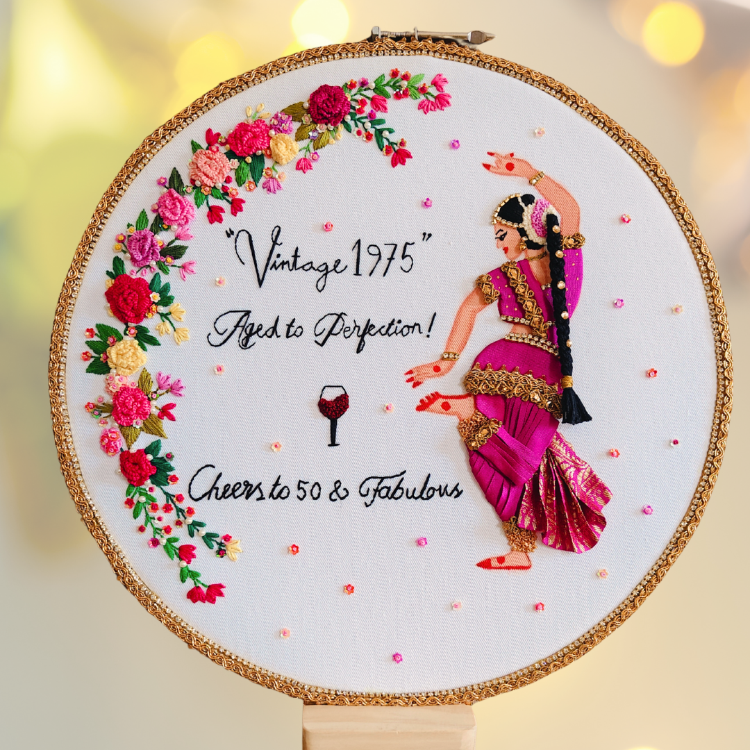 Embroidery hoop with a woman dancing in a pink saree, vintage lettering of cheering 50 years, wine glass embroidered icon, floral decorations, and text on a white background.