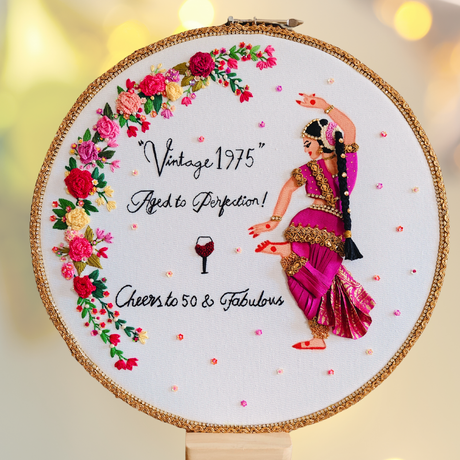 Embroidery hoop with a woman dancing in a pink saree, vintage lettering of cheering 50 years, wine glass embroidered icon, floral decorations, and text on a white background.