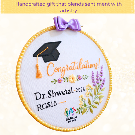 A circular embroidered graduation hoop with a white fabric center, featuring a black graduation cap, orange 'Congratulations!' text, personalized black text reading 'Dr. Shwetal.