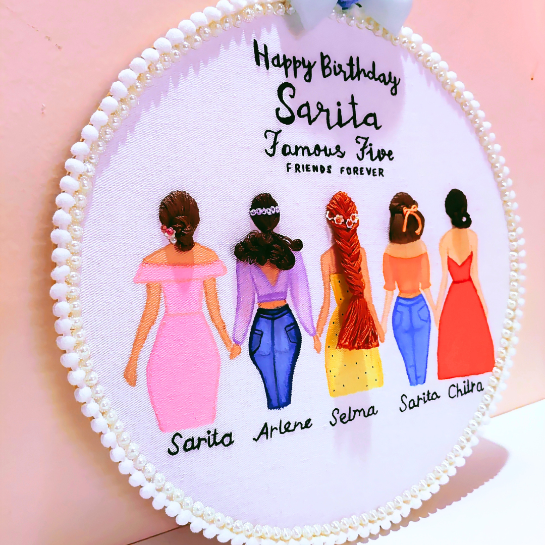 Personalized circular embroidery wall hanging featuring five illustrated girls in colorful dresses with names underneath and the text 'Happy Birthday Sarita Famous Five Friends Forever' on a pale pink background.