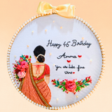 A circular embroidered wall hanging or hoop art piece with a pale blue fabric background, featuring a stitched woman in a red saree from the back, floral motifs, and the text 'Happy 46th Birthday Amma You are like fine wine.' A pale yellow satin bow is attached at the top, and the piece is photographed against a soft peach background.