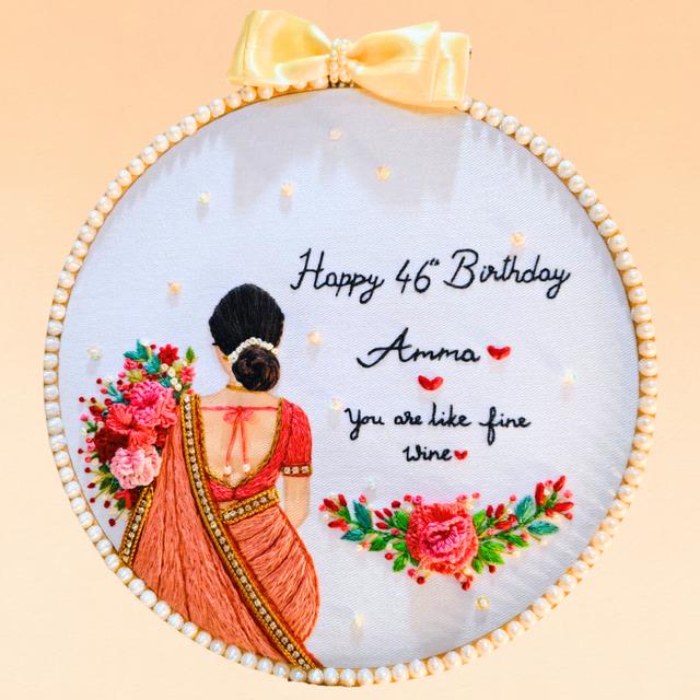 A circular embroidered wall hanging or hoop art piece with a pale blue fabric background, featuring a stitched woman in a red saree from the back, floral motifs, and the text 'Happy 46th Birthday Amma You are like fine wine.' A pale yellow satin bow is attached at the top, and the piece is photographed against a soft peach background.