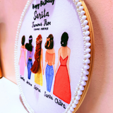 A close-up of a circular embroidered wall hanging in a wooden hoop, featuring illustrated women in colorful dresses with black hair and names beneath them, plus decorative white pom-pom trim around the edge, set against a soft pink background.